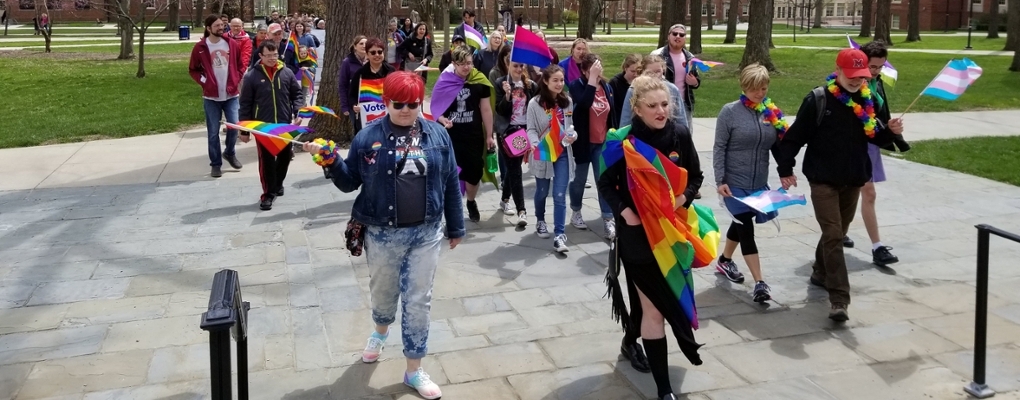 Group of students, faculty and staff holding rainbow flags and walking across academic quad