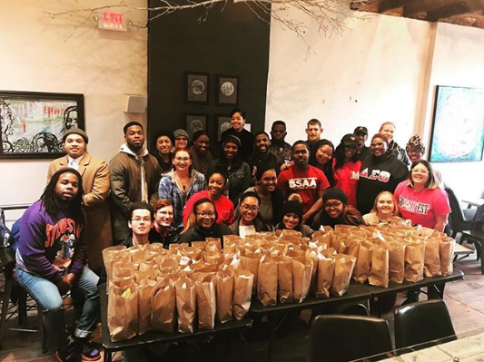 Large group of students posing in front of dozens of lunch bags, recently packed for hashtag lunchbag event.