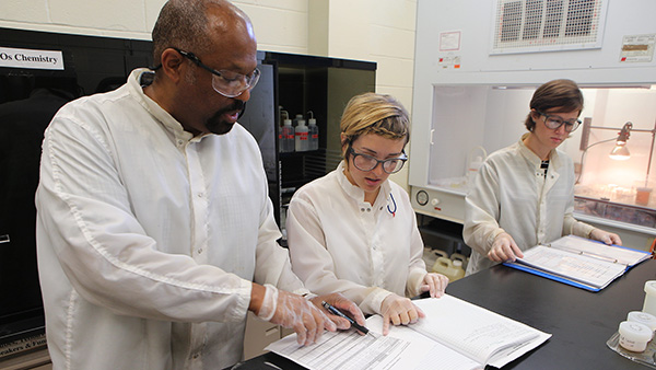  Instructor and two students wearing  protective gear and looking at paperwork in a lab.