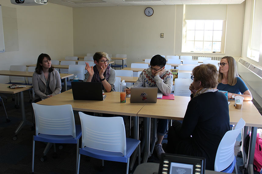 A professor sits at a table with four graduate students in conversation