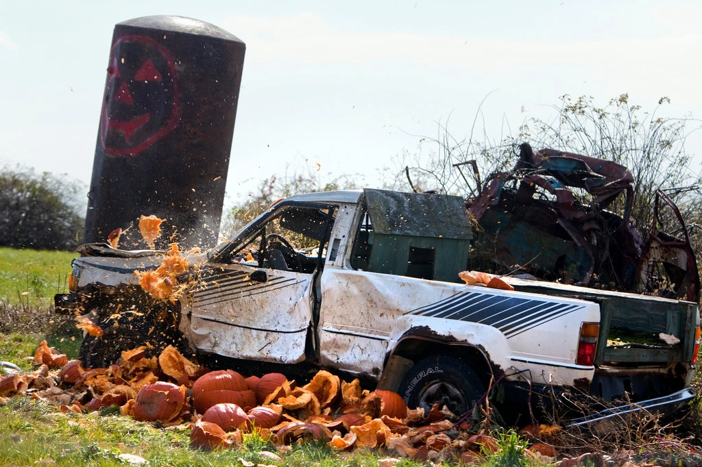 A photo of a battered pick-up truck surrounded by smashed pumpkins. The truck and pumpkins are in the right foreground of an overgrown field and a leaning silo is beyond the truck. Exploding fragments of pumpkin are in the air above the truck as though the truck had just hit the pumpkin or somone had thrown the pumpkin at the truck.