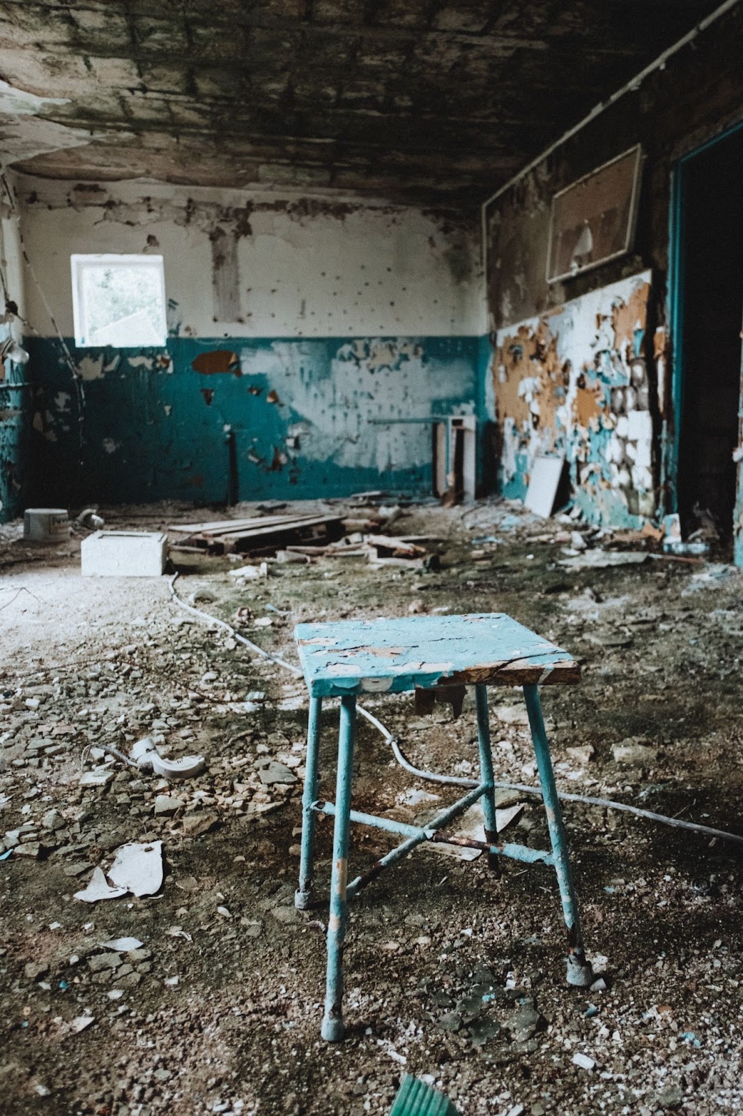 Photo of an abandoned room full of rubble and debris from teh ceiling and crumbling walls. A small blue table is in the foreground and lower half of the far wall is painted blue. The walls are peeling and distressed.