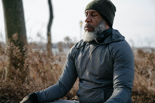 Photo of a man in a jogging suit and kinitted cap, sitting cross-legged in a field of dead grasses.His eyes are closed and he appears to be meditating.