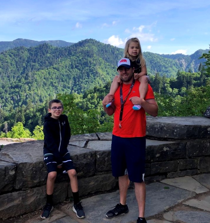 Nick Laes stands with his family in front of a green mountainous landscape. His daughter sits on his shoulders and his son sits by his side