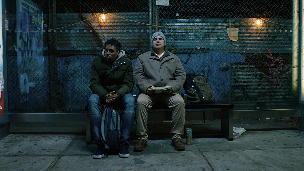 A young man sits on a bus bench next to a DeafBlind man. They are both wearing jackets. The DeafBlind man is wearing a toboggan and holding a notebook.