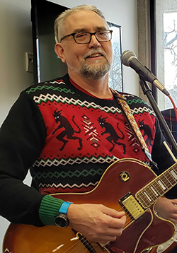Tim Gruenhagen, a white man with glasses and gray hair, wearing a festive sweater and holding a guitar