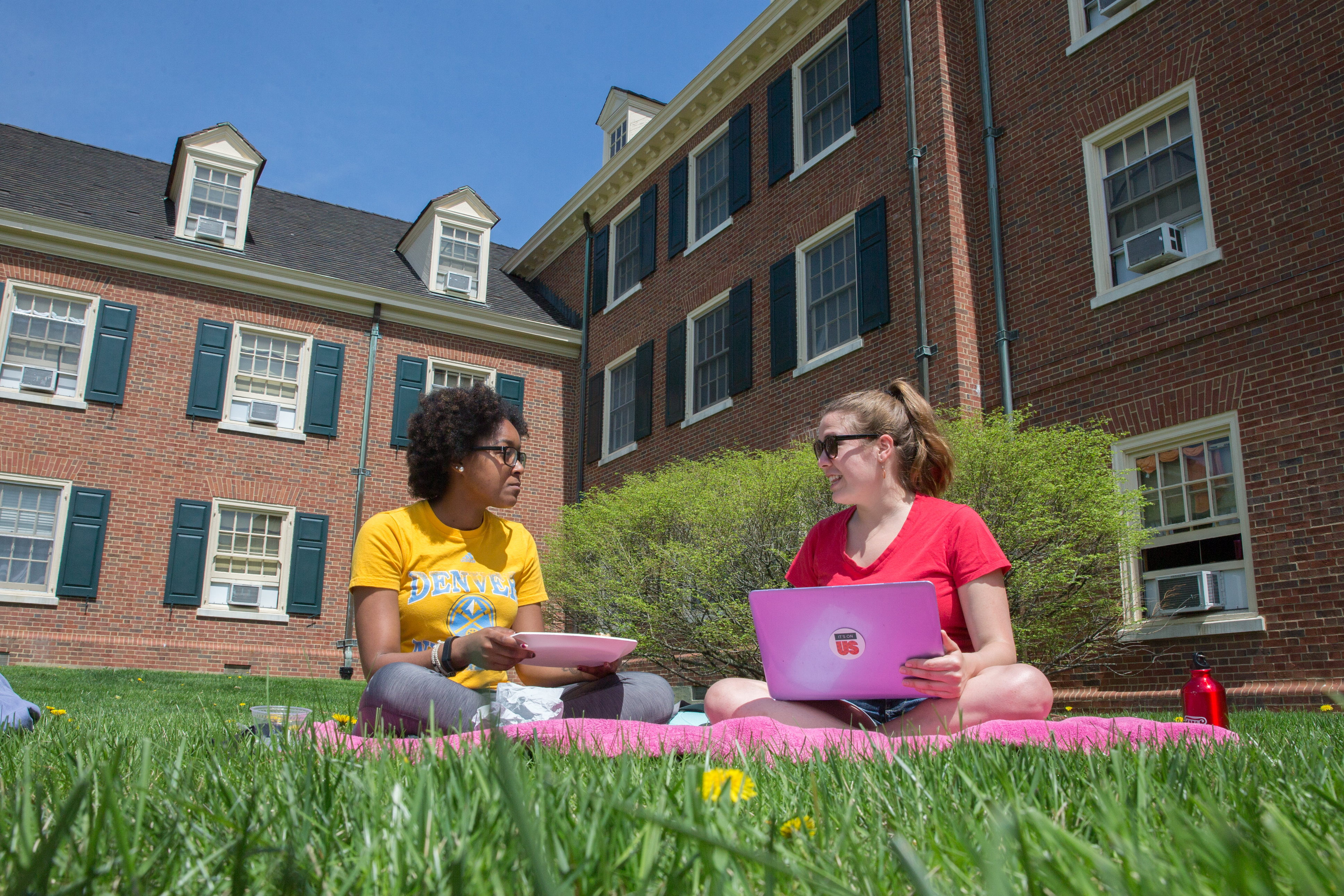 Two female students with laptops sitting on a blanket outside their residence hall