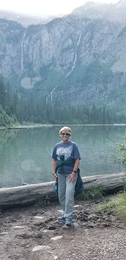 Roxanne Storer, a brown-haired woman, stands in front of a river in hiking boots, smiling at the camera