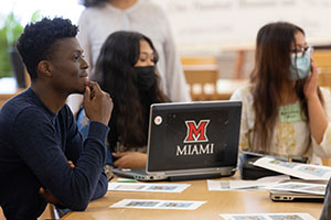 Students wearing masks sitting around a computer with the Miami M logo