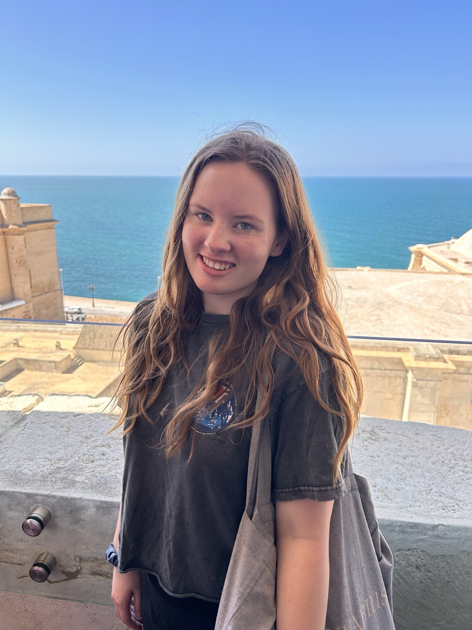 Photo of young caucasian woman (Xandra) standing on a structure in front of the ocean. She has long golden brown hair and blue eyes. She is wearing a dark grey t-shirt and has a grey tote bag on her shoulder.