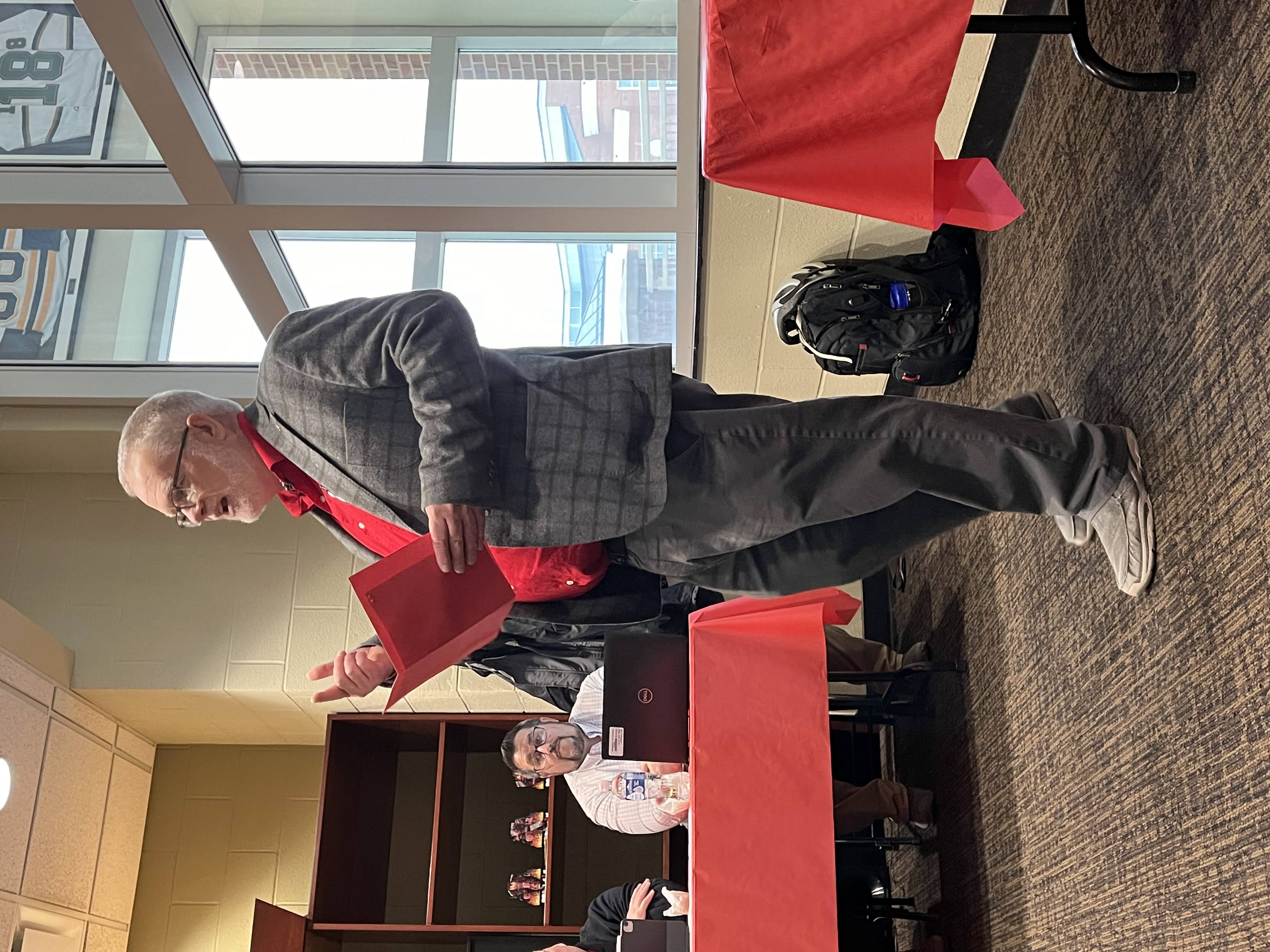 Tim Jones, wearing a red Oxford shirt and sport coat, stands in front of a table of judges, emceeing