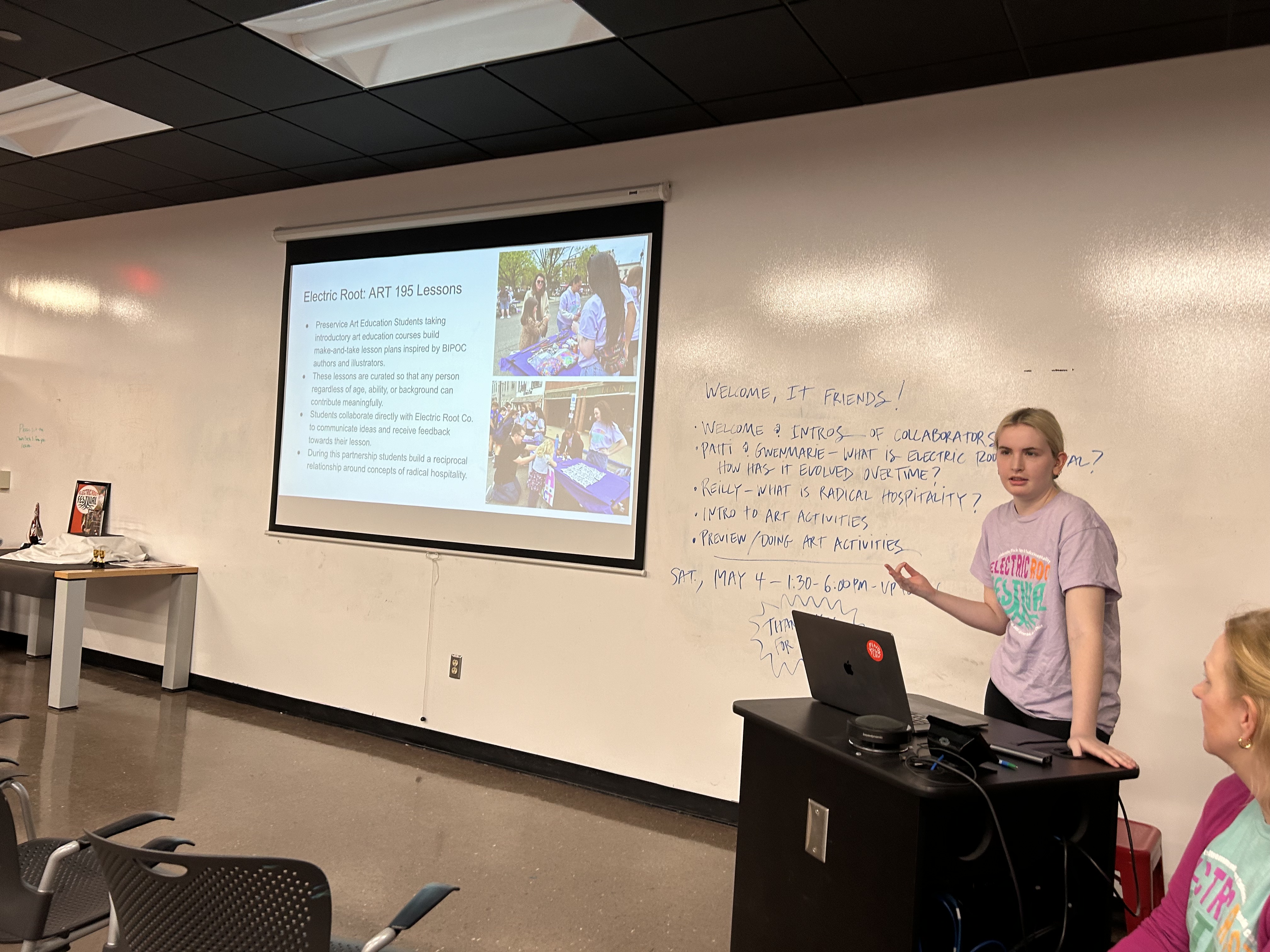 Reilly Powers, a femme-presenting person with long blonde hair and a purple shirt, stands at a classroom podium giving a presentation about radical hospitality