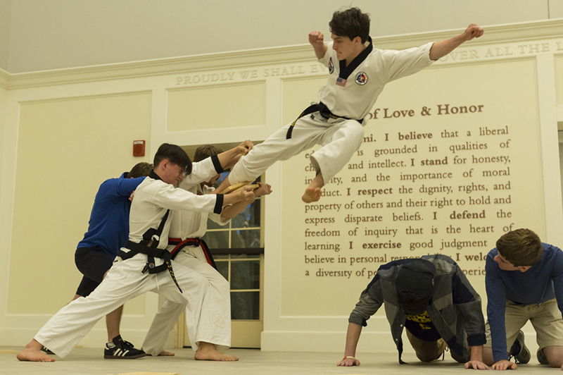 A blackbelt jumps over two students on their hands and knees and breaks a board in a Taekwondo demonstration in the Shade Family Room