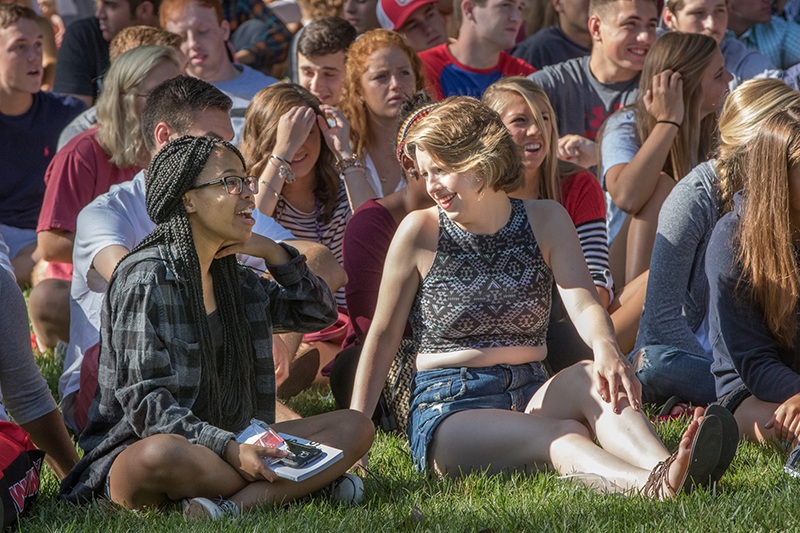 Two students talk to each other while sitting in a crowd on the grass