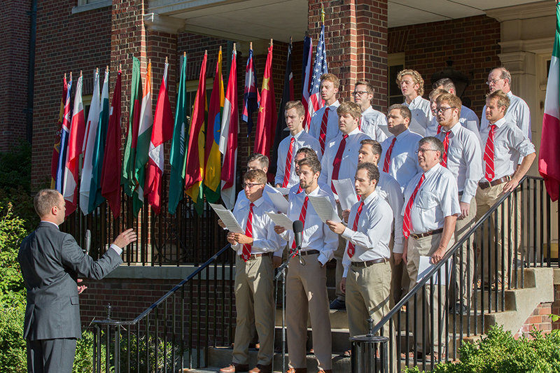 A men's chorus sings beside a line of international flags