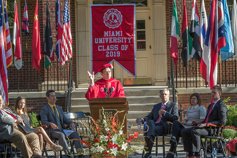 President Hodge gestures as he speaks at convocation. Behind him is a line of international flags and a Miami University class of 2019 banner