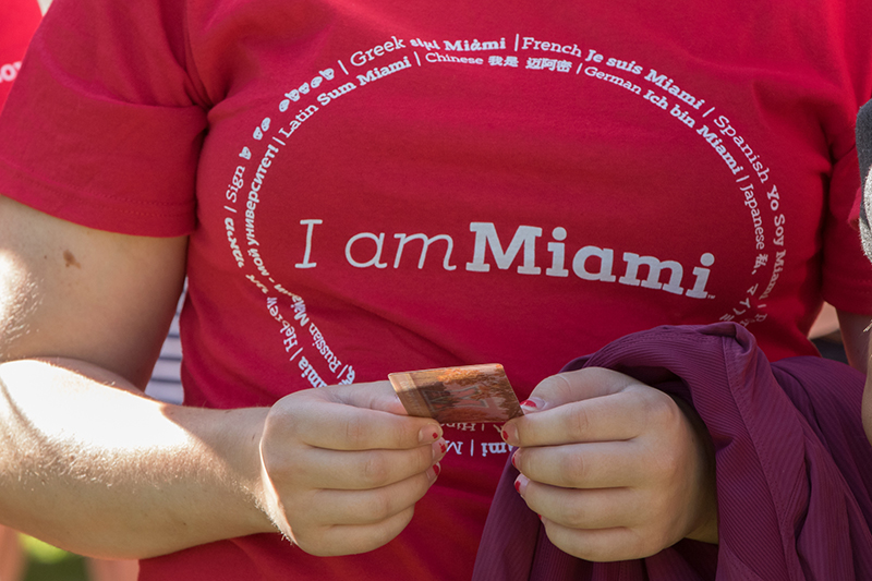 A student wearing an I am Miami shirt and holding a card with a photo of central quad