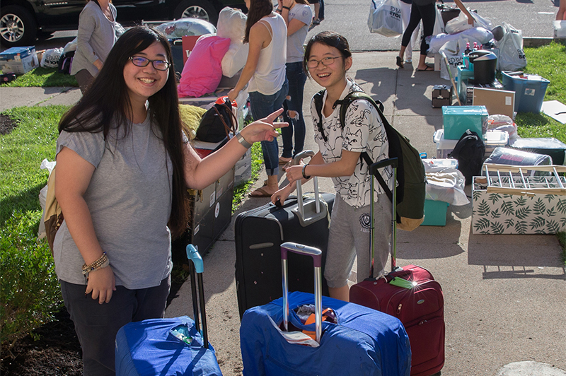 Two female students smile and stand by their many suitcases