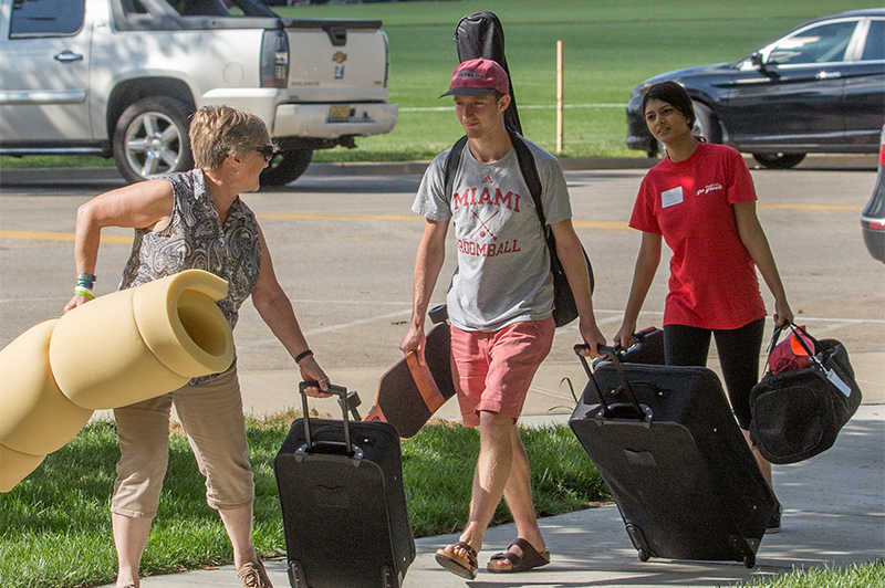 A student, a parent, and a volunteer carry a mattress topper, a guitar, and suitcases down a sidewalk