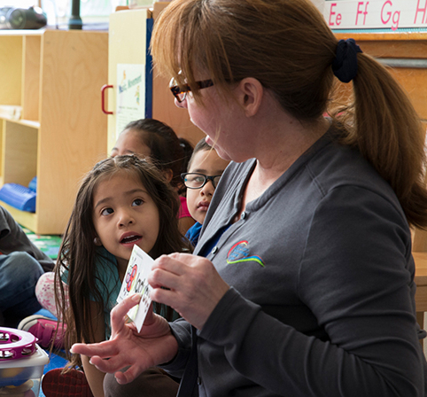 Young English language learners in a classroom (pre-COVID-19) work with their instructor using flashcards.