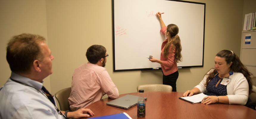 Student writing on a dry-erase board in a meeting with three other people