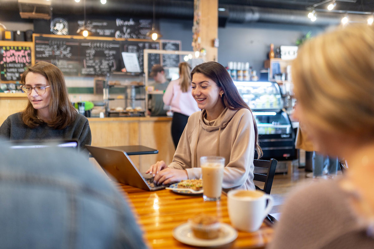 A smiling student looking at their laptop while sitting in at a coffee shop table