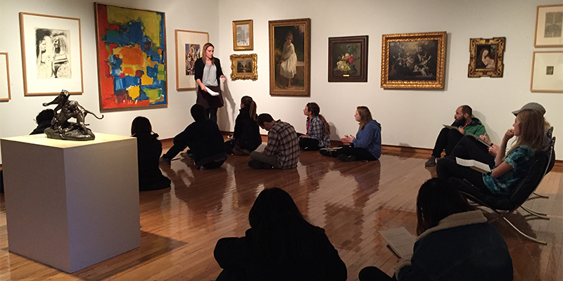 A group of students seated in a gallery as a professor speaks before a group of paintings