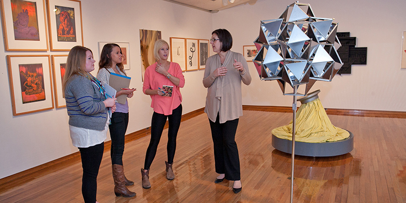 A group of students listen as a professor speaks in front of a silver geometric sculpture 