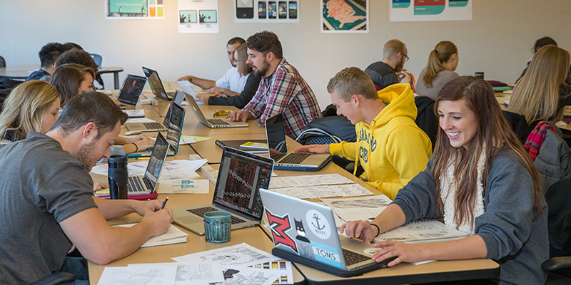  Students seated at rectangular tables work on computers during a typography class