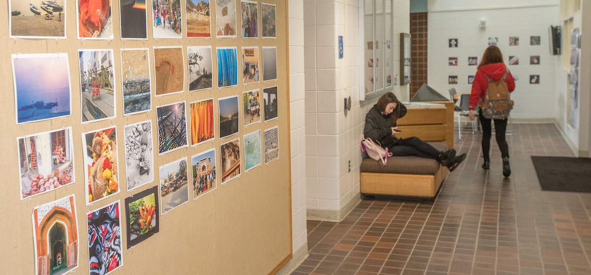  Prints are pinned to an Art Building bulletin board. Students work and walk in the hallway.