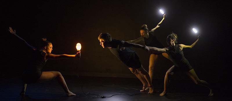 On a darkened stage, dancers pose with lights in their hands
