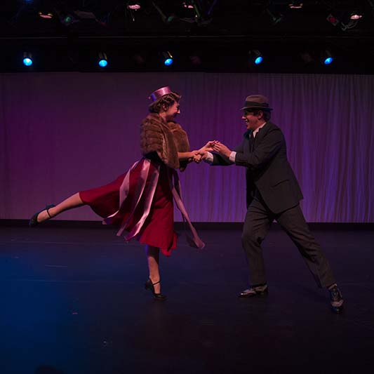 A dancing couple dressed in 40s fashion face each other, holding hands