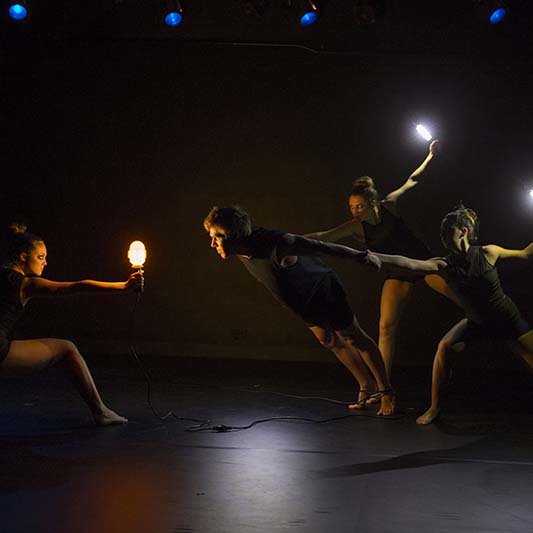 On a darkened stage, dancers perform while holding illuminated devices