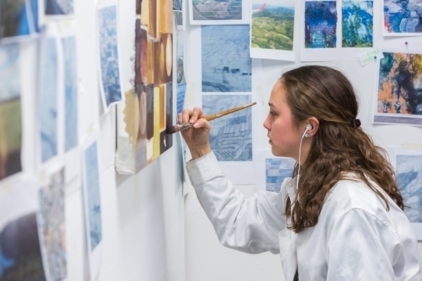 A student works on a painting in the studio, surrounded by colorful scenes tacked to the walls.