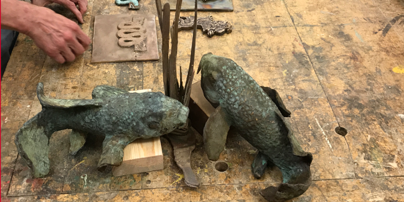 An artist's hand is visible near two bronze fish on a worktable
