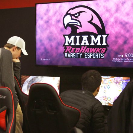 The Miami Redhawks Varsity Esports logo is illuminated on a large screen. In foreground, students gaze at computer screens.