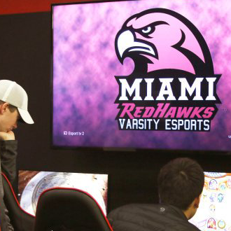 Seen from behind, students working on computers in the Miami esports arena. A Redhawks varsity esports logo is visible on the wall.