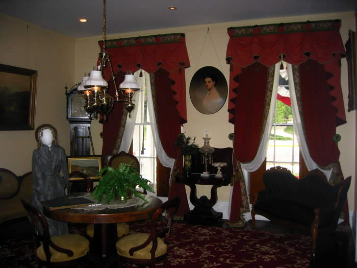 Parlor, with furnishings including drapery, lamps, portraits, a round table, and chairs. A mannequin is dressed in the style of a 19th century woman.