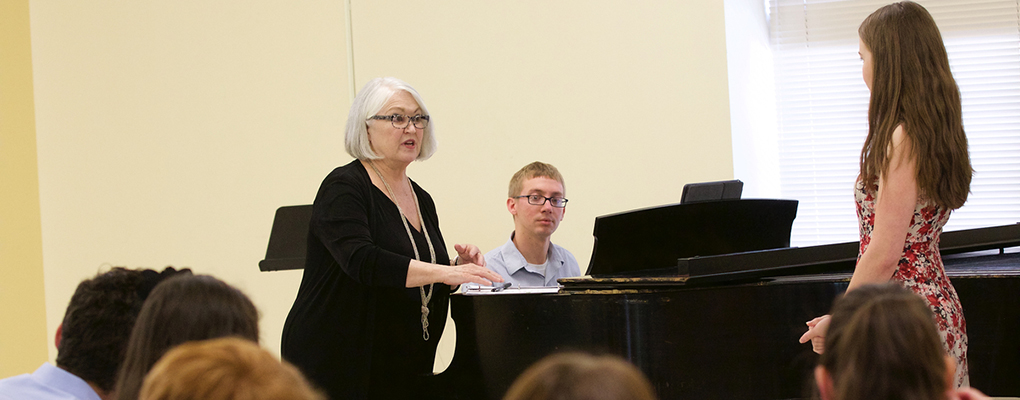 An instructor stands near the piano and offers advice to a vocalist
