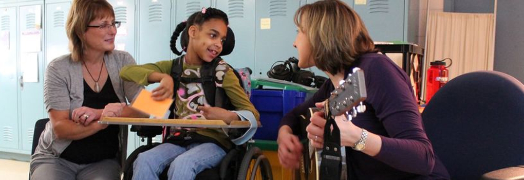 A music therapist plays guitar for a smiling child as another woman looks on