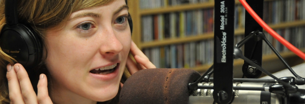 Woman holding her fingers to the sides of her headphones speaks into a broadcast microphone 
