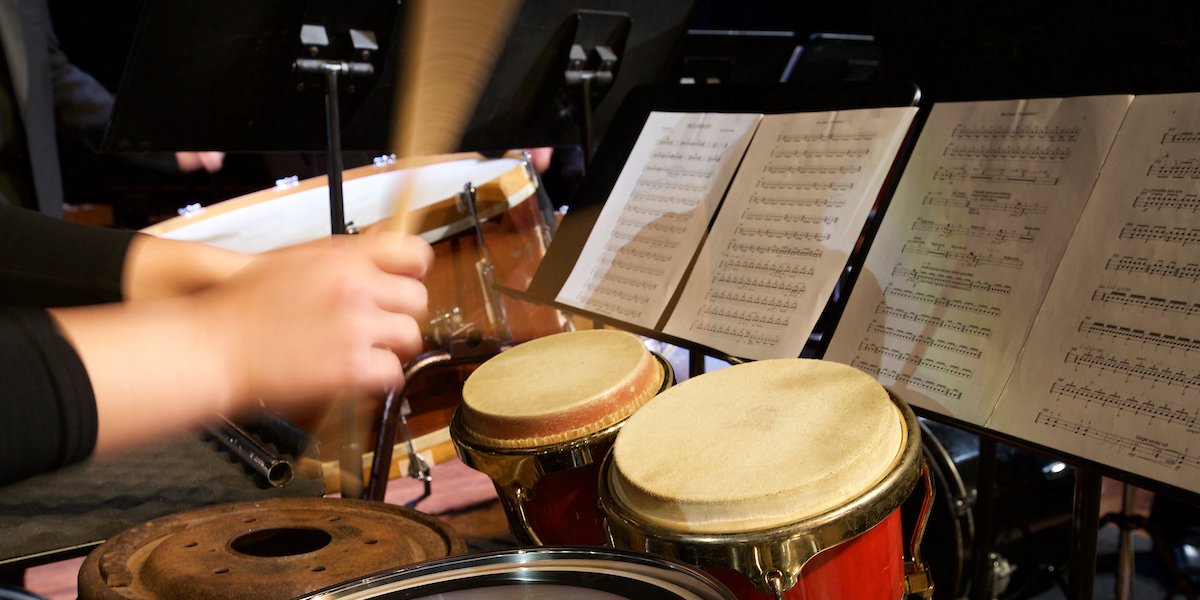 With music on a stand, hands holding sticks are striking drums in a kit that includes world percussion instruments at a concert