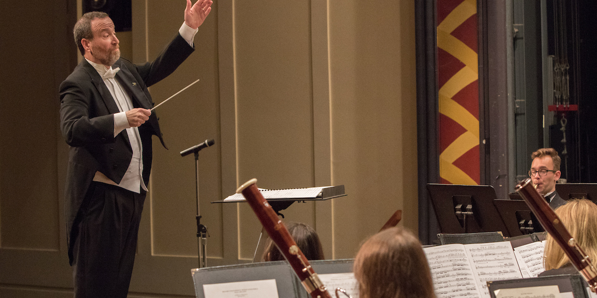 As seen from within the ensemble, the conductor lifts his hand to give a cue. Bassoonists are visible in front.