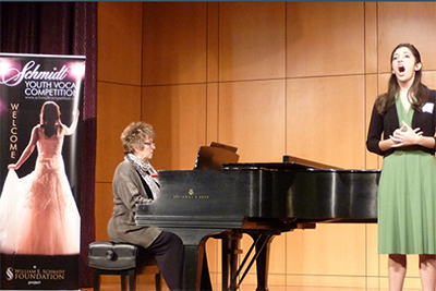 A young woman sings onstage, accompanied by a pianist. Banner at left 'Schmidt Youth Vocal Competition'