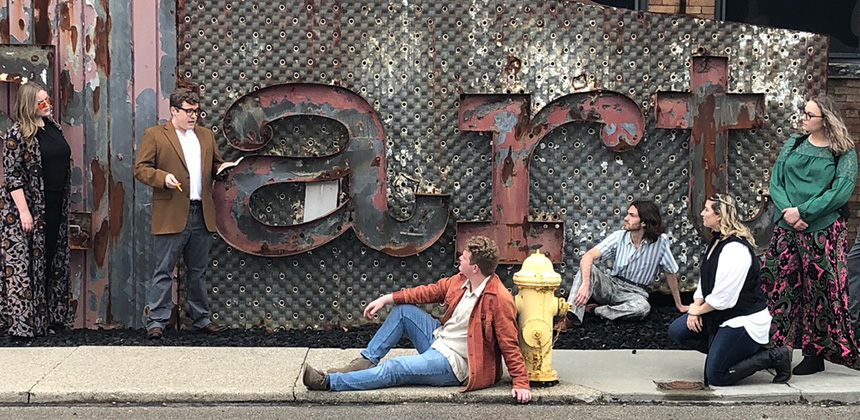 Young men and women dressed in sixty's era clothes lounging in front of an old and rusty sign that reads "art"