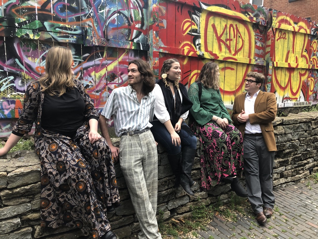  Actors three women and two men in sixties era cloths having a conversation in front of a graffiti covered wall