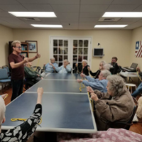 Residents of Knolls of Oxford each hold a length of rope, seated around a large table as Kevin Spencer teaches a magic trick