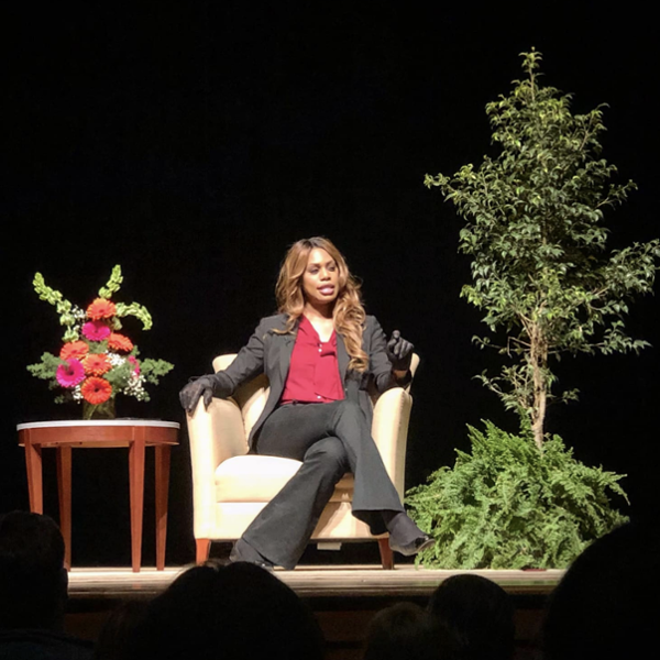 Laverne Cox sits in an easy chair onstage, flanked by plants and a small table