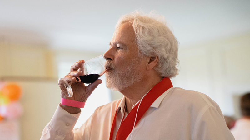  A man with wavy white hair and  white beard brings a glass of red wine to his lips to taste