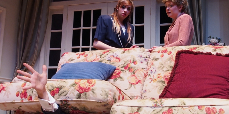 A hand reaches from beneath the couch cushions as two women look on in a scene from Communicating Doors